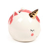 Angled view of a white ceramic unicorn money box, featuring a shiny gold horn, pink inner ears, pink mane, and pink cheek details. Its eyes are closed black lines, and the coin slot is visible on top.