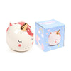 Angled view of a white ceramic unicorn money box with a golden horn, pink ears, cheeks, and mane, featuring closed eyes, alongside its blue patterned presentation box.