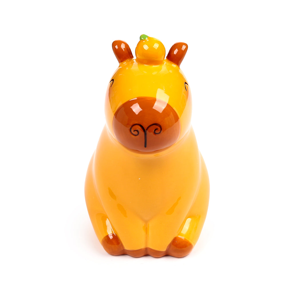 Front view of a glossy, yellow and brown ceramic capybara money box, with small black eyes, a dark brown nose featuring swirly nostrils, brown ears, and a small green-topped yellow fruit on its head.
