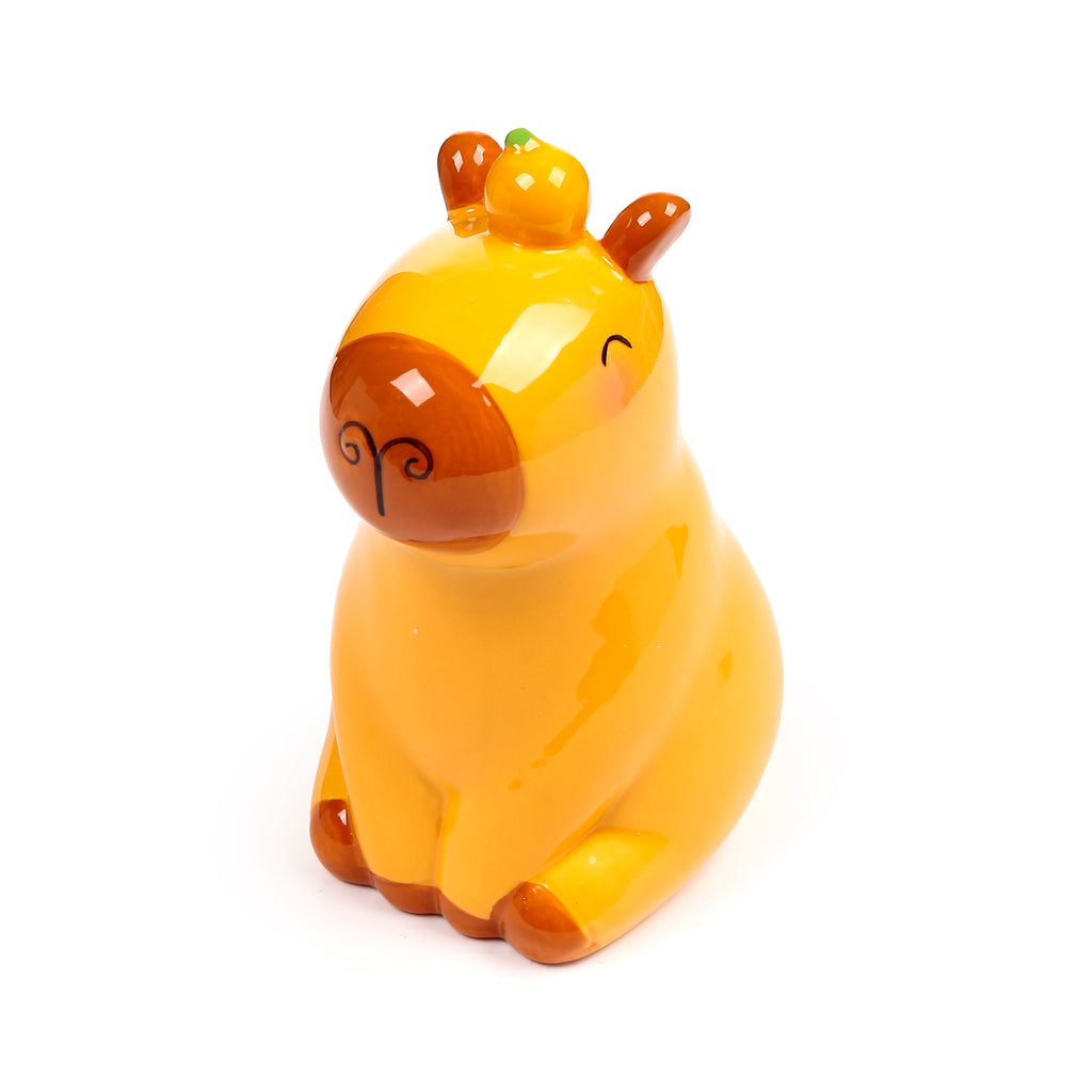Angled view of a glossy yellow-orange ceramic capybara money box with brown snout, ears, and feet, and a small orange fruit with a green stem on its head.