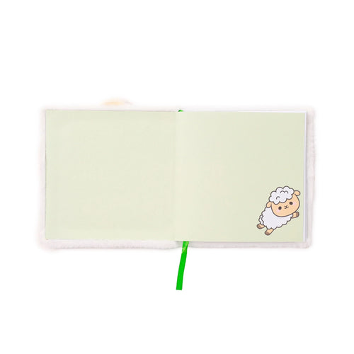 Overhead view of an open plush notebook with a white fluffy cover, featuring light green pages and a bright green ribbon bookmark, with a cute cartoon sheep illustration on the right-hand page.