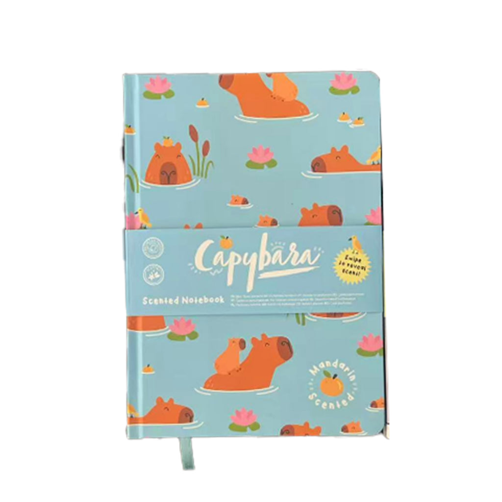 Front view of an A5 notebook with a light blue cover depicting cartoon capybaras swimming amongst pink lily pads and green reeds. A paper band across the front identifies it as a 'Capybara Scented Notebook' with 'Mandarin Scented' text. A light blue ribbon bookmark is visible.