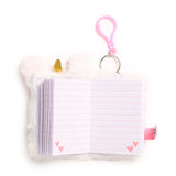 Overhead view of an open white plush unicorn notebook with a gold glitter horn. Its lined white pages feature pink hearts at the bottom corners. A pink glitter pen is attached to the notebook via a metal keyring and pink carabiner clip.