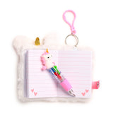 Top-down view of an open white plush A7 notebook with a golden unicorn horn, pink lined pages featuring hearts, and a pink multi-colour unicorn pen. A pink carabiner keyring is attached.