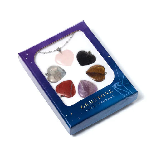 Angled view of a dark blue boxed set, labelled 'GEMSTONE HEART PENDANT', showcasing six interchangeable heart-shaped gemstone pendants. A pink heart pendant is attached to a silver-coloured chain, displayed with additional black, brown, red, purple, and grey heart gemstones.
