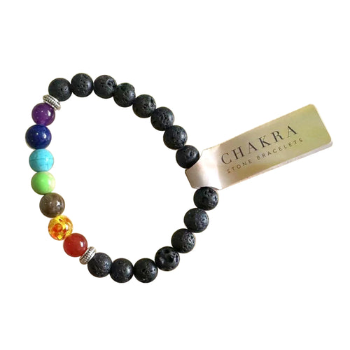 Overhead view of a chakra lava rock gemstone bracelet featuring porous black lava beads, smooth, colourful chakra gemstone beads (purple, dark blue, turquoise, green, brown, orange, red), and silver-coloured spacer beads. A light-coloured tag reading 'Chakra Stone Bracelets' is attached.