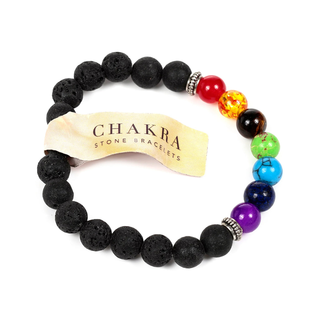 Angled view of a coiled chakra lava rock gemstone bracelet, featuring black porous lava beads, seven polished gemstone beads in chakra colours (red, orange, brown, green, light blue, dark blue, purple), and ornate silver-coloured spacers. A light-coloured tag reading 'CHAKRA STONE BRACELETS' is attached to the bracelet.