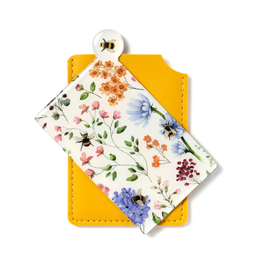 Overhead, angled view of a pocket mirror with a white surface, depicting a nectar meadows bee and colourful floral pattern, partially inserted into a bright yellow protective sleeve.