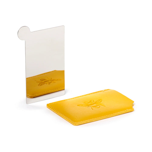 Angled view showing a silver-coloured rectangular pocket mirror standing upright, next to a yellow protective sleeve with an embossed bee design, reflected in the mirror.