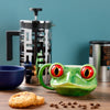 Lifestyle shot of a green ceramic tree frog head shaped mug with prominent red and black eyes, set on a wooden table with a silver cafetière, a blue bowl, scattered coffee beans, and biscuits.