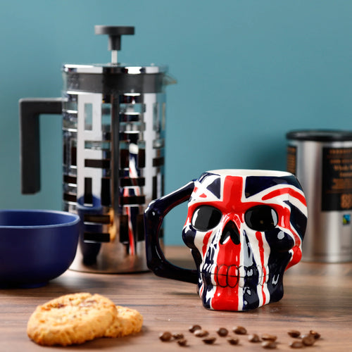 Front view of a UK Flag Skull Head Ceramic Shaped Mug in a lifestyle setting. The mug features a red, white, and blue Union Jack design on the skull head, with black eye sockets and a dark handle. It is on a wooden table with biscuits and coffee beans scattered nearby.