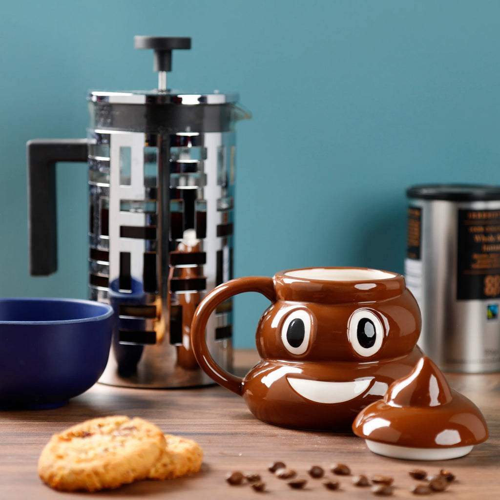 Front view of a brown ceramic poop-shaped mug with a white smiling face and black eyes, its matching brown lid detached. The mug sits on a wooden surface surrounded by cookies, scattered coffee beans, and a blurred French press against a blue background.