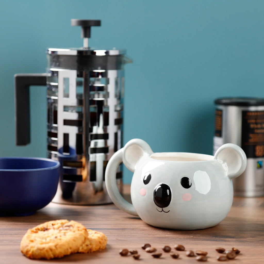 Angled lifestyle shot of a light grey ceramic koala mug, showing its shaped head with black eyes, nose, and pink cheeks, with a handle on the right. It sits on a wooden surface beside biscuits and scattered coffee beans.