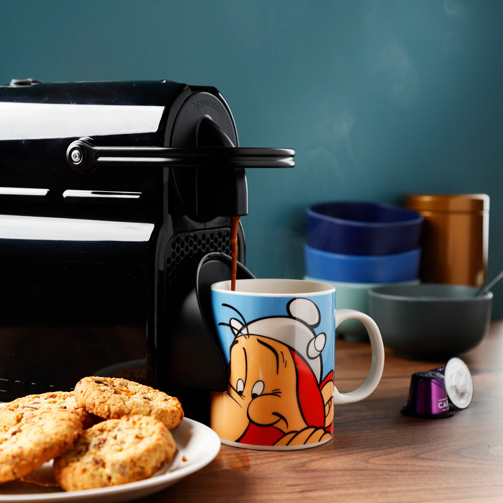 Lifestyle shot of an Asterix Obelix porcelain mug, light blue with a white handle and a cartoon depiction of Obelix, being filled with coffee from a machine. Steam rises from the mug, with biscuits and coffee capsules on the wooden surface, and colourful bowls in the background.