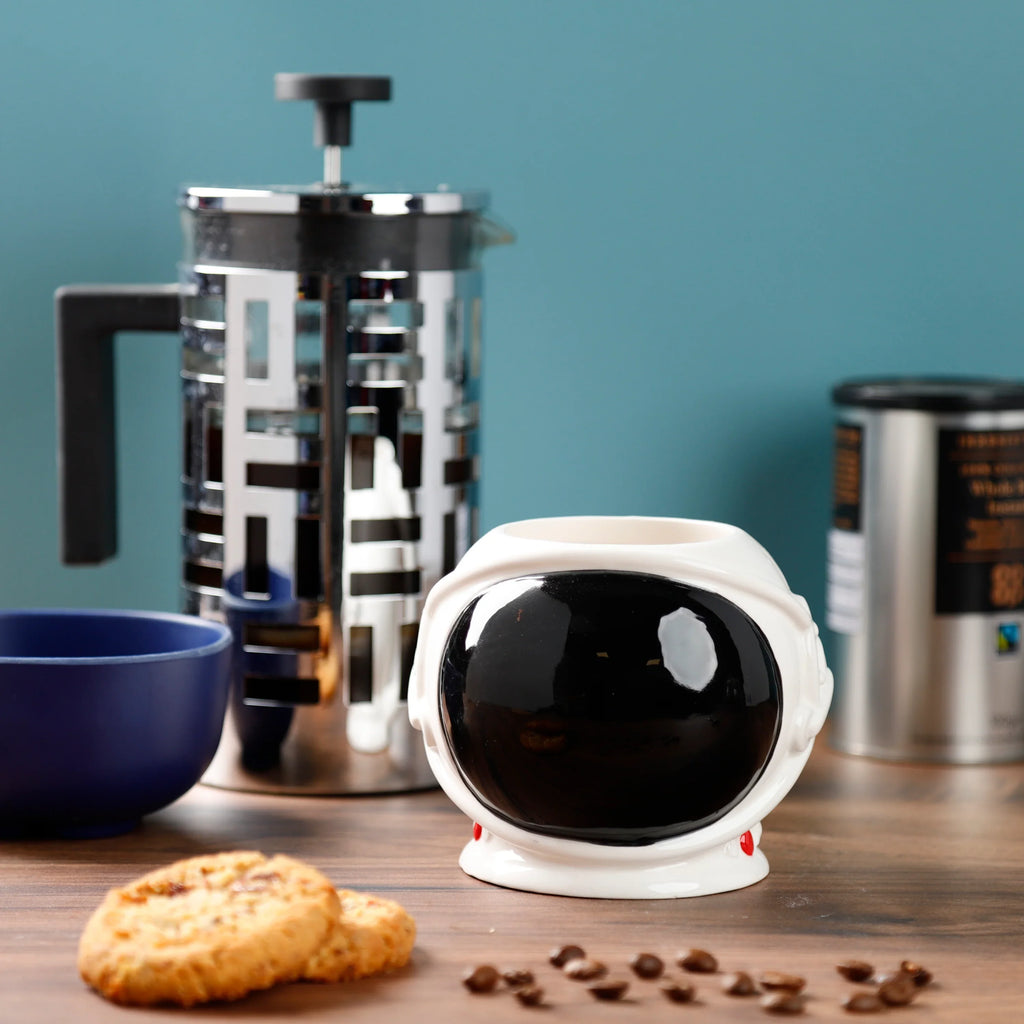 White ceramic astronaut space helmet mug with a large black visor, seen from a front-angled view on a wooden surface, surrounded by coffee beans, cookies, a French press, and a blue bowl.