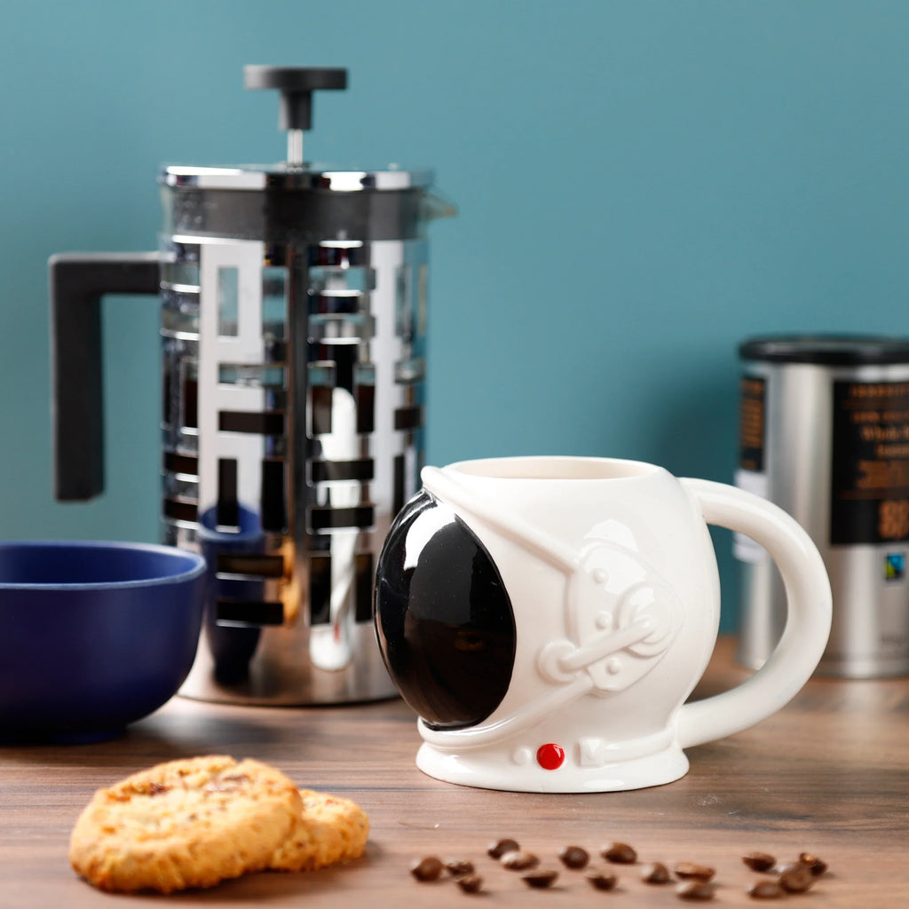 Angled lifestyle shot of a white ceramic astronaut's helmet mug with a black visor and red button, on a wooden surface with coffee beans, cookies, and coffee brewing equipment.