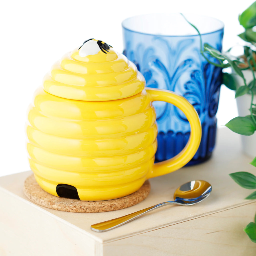 Lifestyle shot of a yellow beehive-shaped ceramic mug, with a lid topped by a bee decoration and a black entrance hole near the base. The mug sits on a cork coaster, alongside a golden spoon, a blue patterned glass, and a green plant.