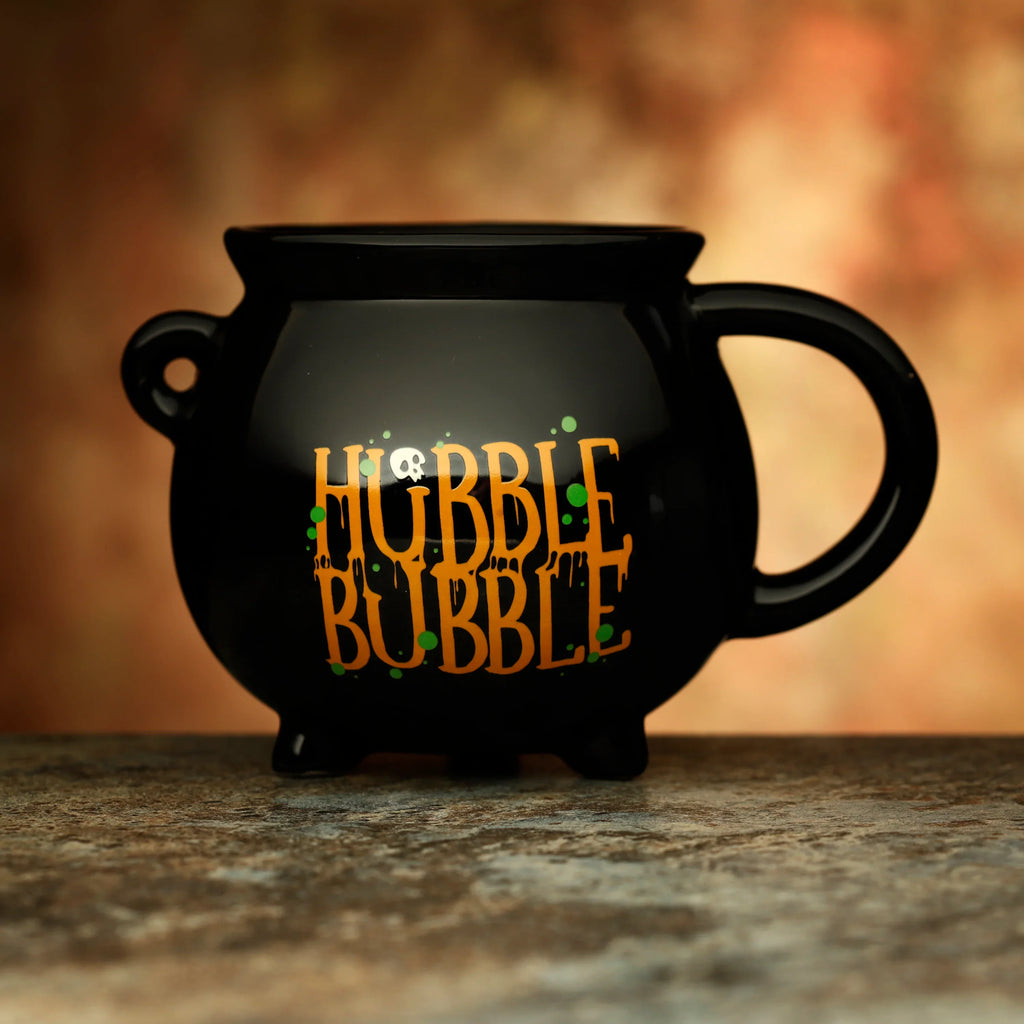 Front view of a black ceramic cauldron mug with orange 'HUBBLE BUBBLE' text, a white skull, and green potion splatters, resting on a dark textured surface.