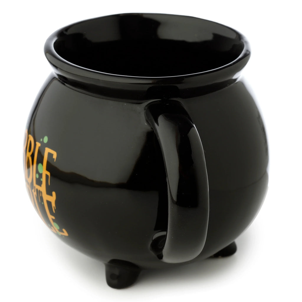 Angled view of a glossy black ceramic cauldron-shaped mug, showing its side handle, partial orange text with green splatters, and three small feet.