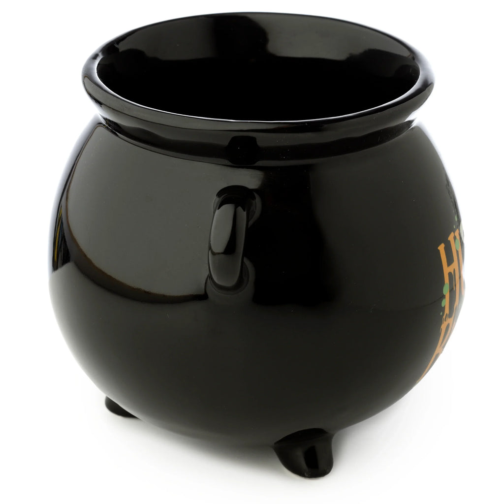Angled view of a glossy black ceramic cauldron-shaped mug, showing its handle, two visible feet, and a partial orange and green decorative design.