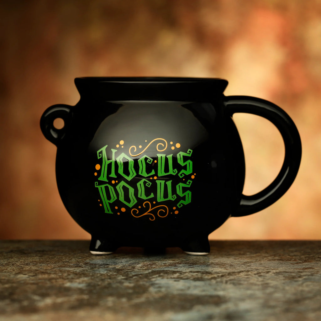 Black ceramic cauldron-shaped mug with green 'Hocus Pocus' lettering, orange decorative accents, and a side handle.