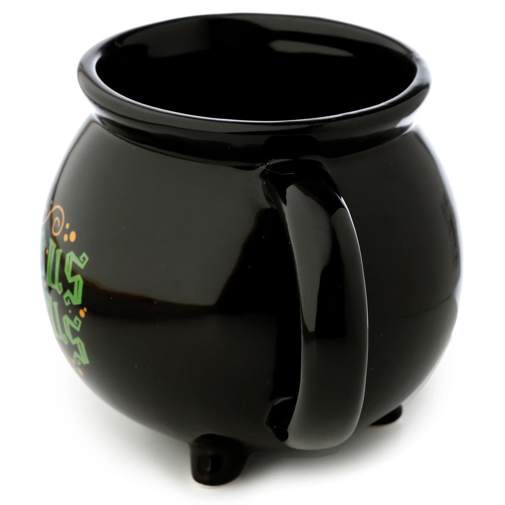 Angled view of a glossy black ceramic cauldron-shaped mug, showing its handle, three small feet, and green stylised lettering with orange accents on the side.