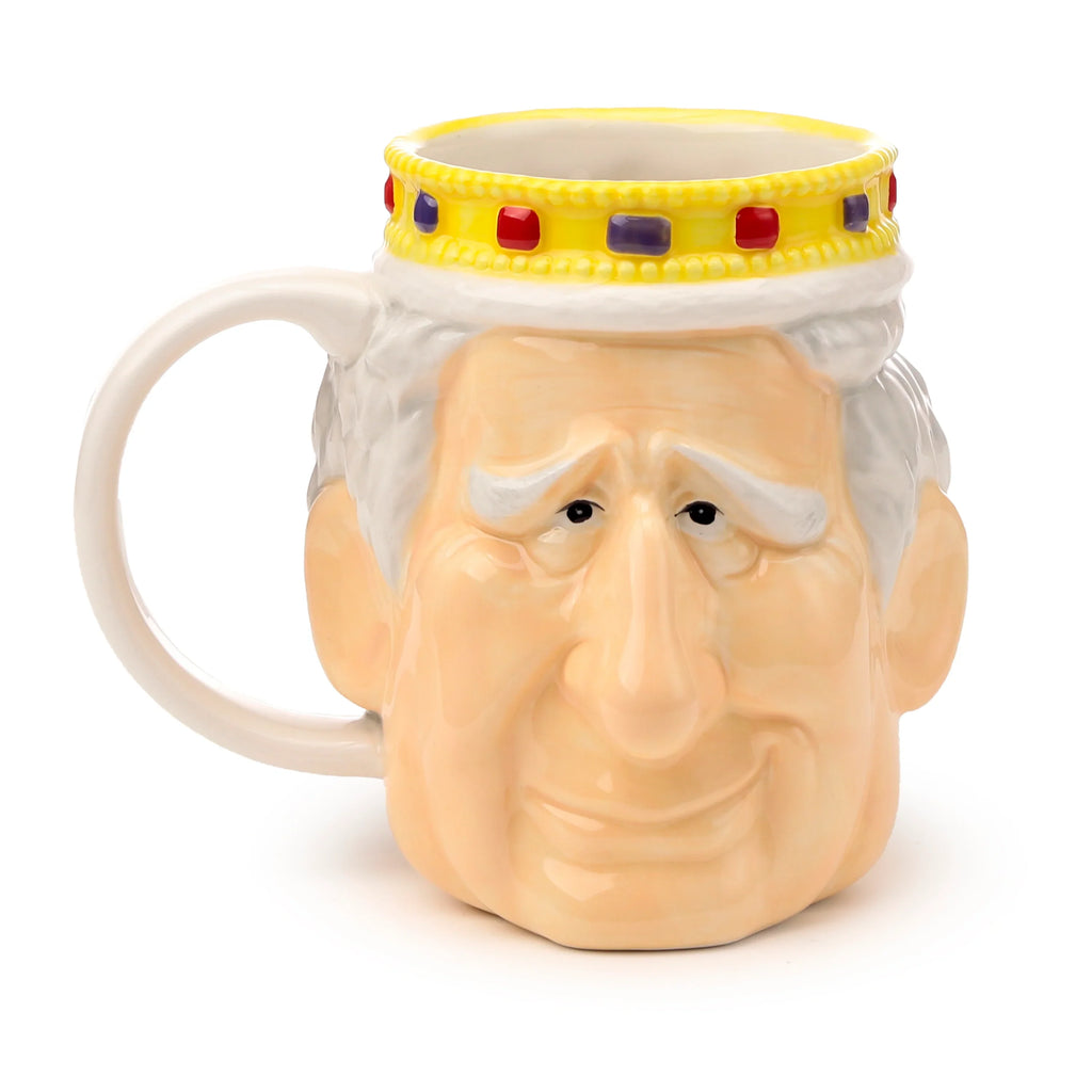 Front view of a ceramic shaped head mug, depicting King Charles III. The light-coloured face features white hair and eyebrows, topped with a yellow crown adorned with red and purple jewels. A cream-coloured handle is visible on the left.