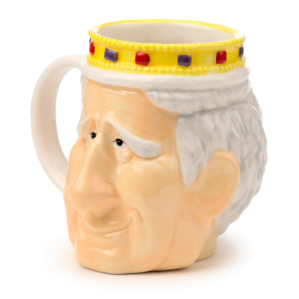 Angled view of a ceramic King Charles III shaped head mug, featuring white hair, a light complexion, and a yellow crown band adorned with red and purple jewels. A cream-coloured handle is visible.