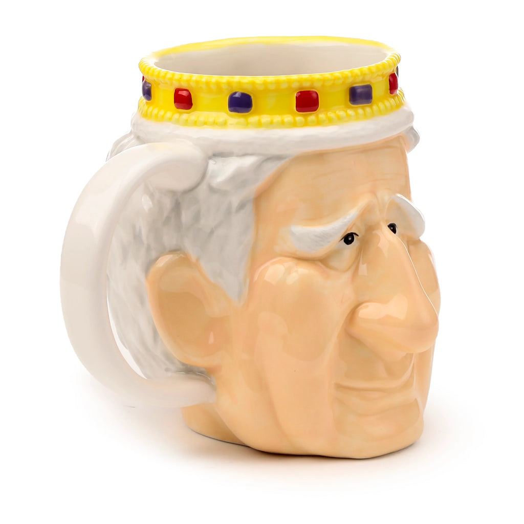 Angled view of a ceramic King Charles III shaped head mug. Features a peach-coloured face with white hair, black eyes, and a yellow crown rim decorated with red and purple square jewels. A white handle is visible.