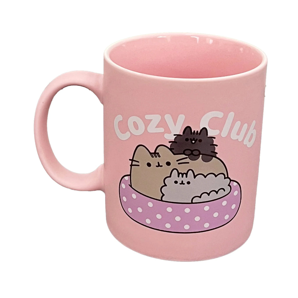 Angled view of a pink porcelain mug, featuring a graphic design of Pusheen the Cat and two smaller cats snuggled in a pink polka-dot bed, with Cozy Club text above.