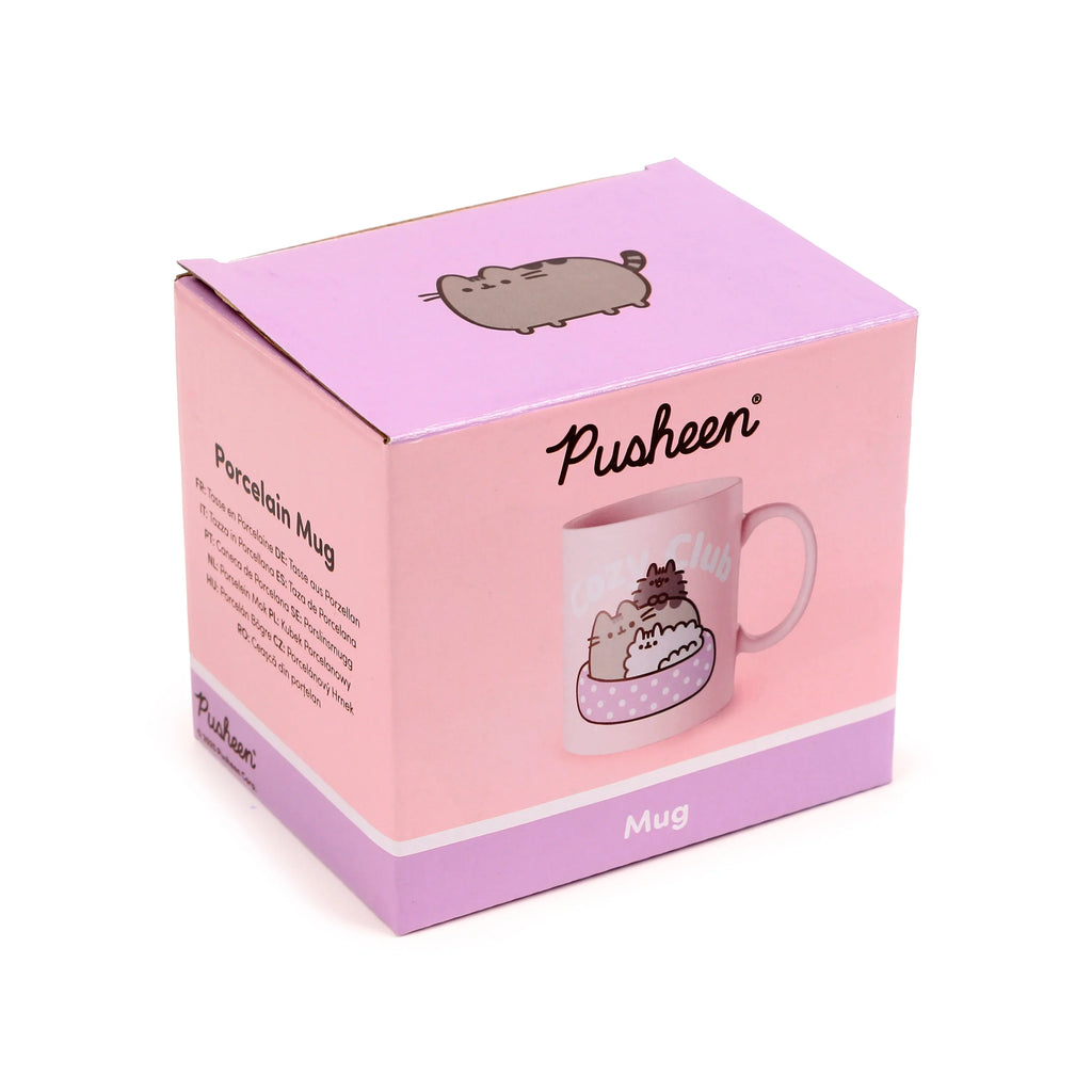 Angled front view of a pink and purple cardboard box, packaging for a porcelain mug, decorated with Pusheen the cat illustrations and 'COZY Club' text.
