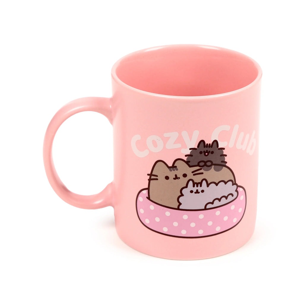 Angled view of a pink porcelain mug, displaying the handle and a 'Cozy Club' illustration featuring three stylised cats snuggled in a pink polka-dot basket.