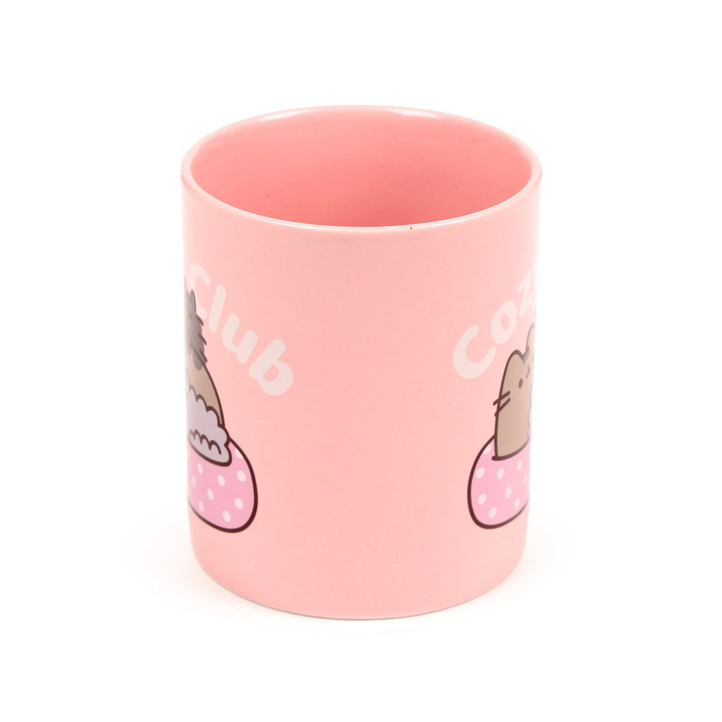 Angled view of a pink porcelain mug, featuring the character Pusheen the Cat snuggled in a pink polka-dot blanket, with partial 'Cozy Club' text visible on the side.