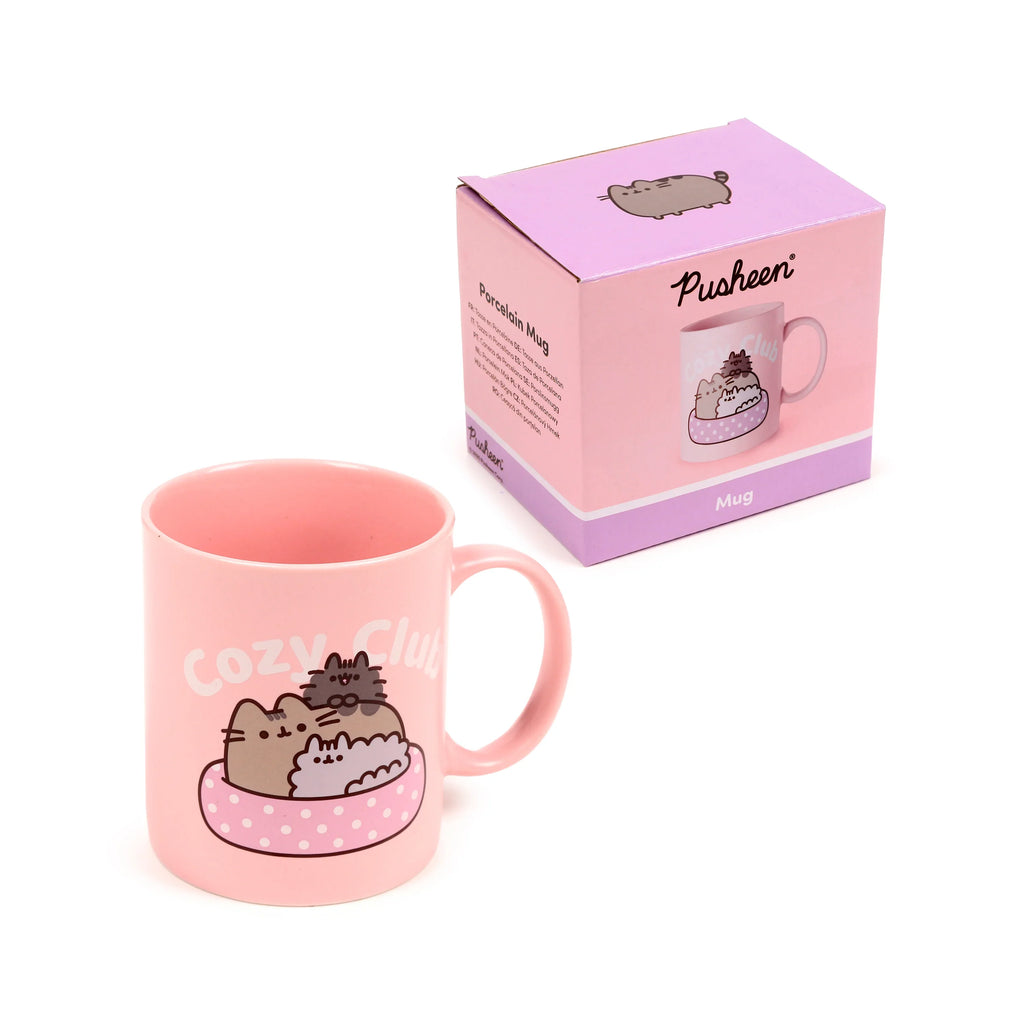Angled view of a pink porcelain Pusheen the Cat mug, featuring two cats snuggled in a polka-dot bed and 'Cozy Club' text, alongside its pink and lavender gift box.