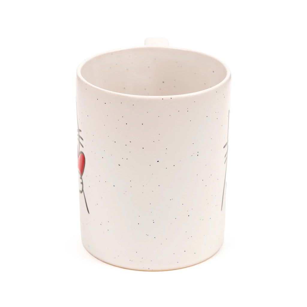 Angled view of a cream speckled stoneware mug, showing a red heart and black line details of a cat design on its side.