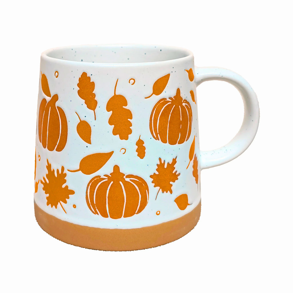Angled view of a white speckled stoneware pumpkin mug, featuring orange pumpkin and autumn leaf patterns, with a terracotta-coloured base and visible handle.