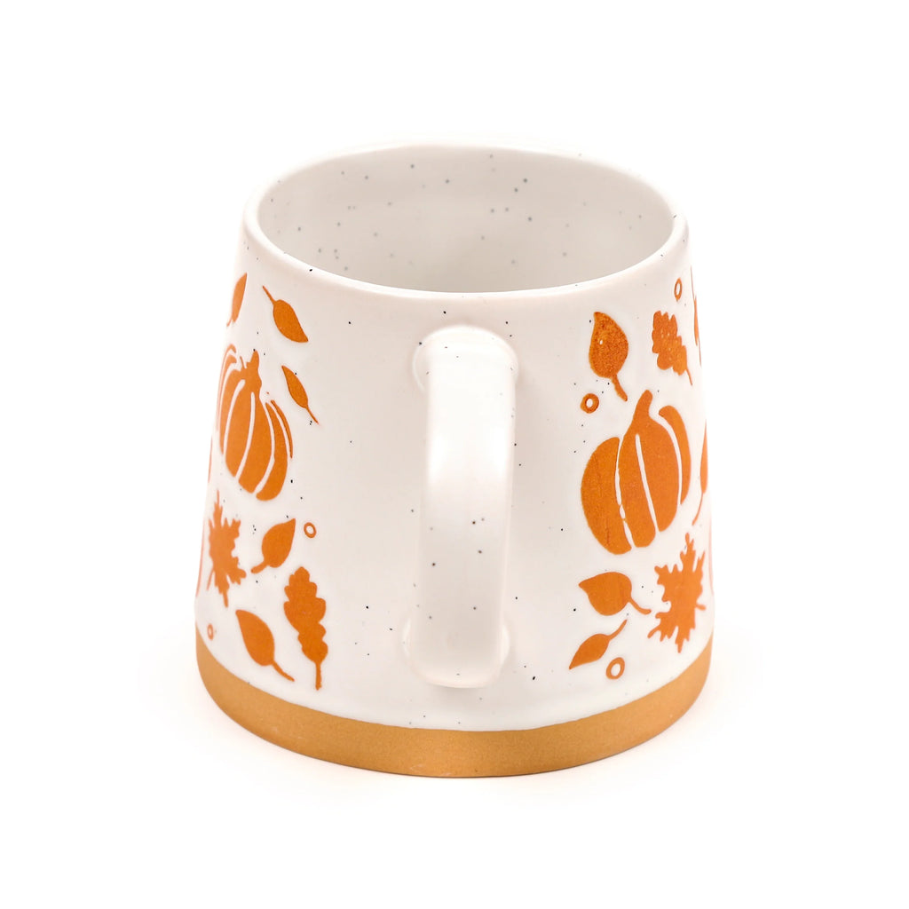 Angled view of a speckled white stoneware mug with orange pumpkin and autumn leaf patterns, showing its handle and unglazed base.