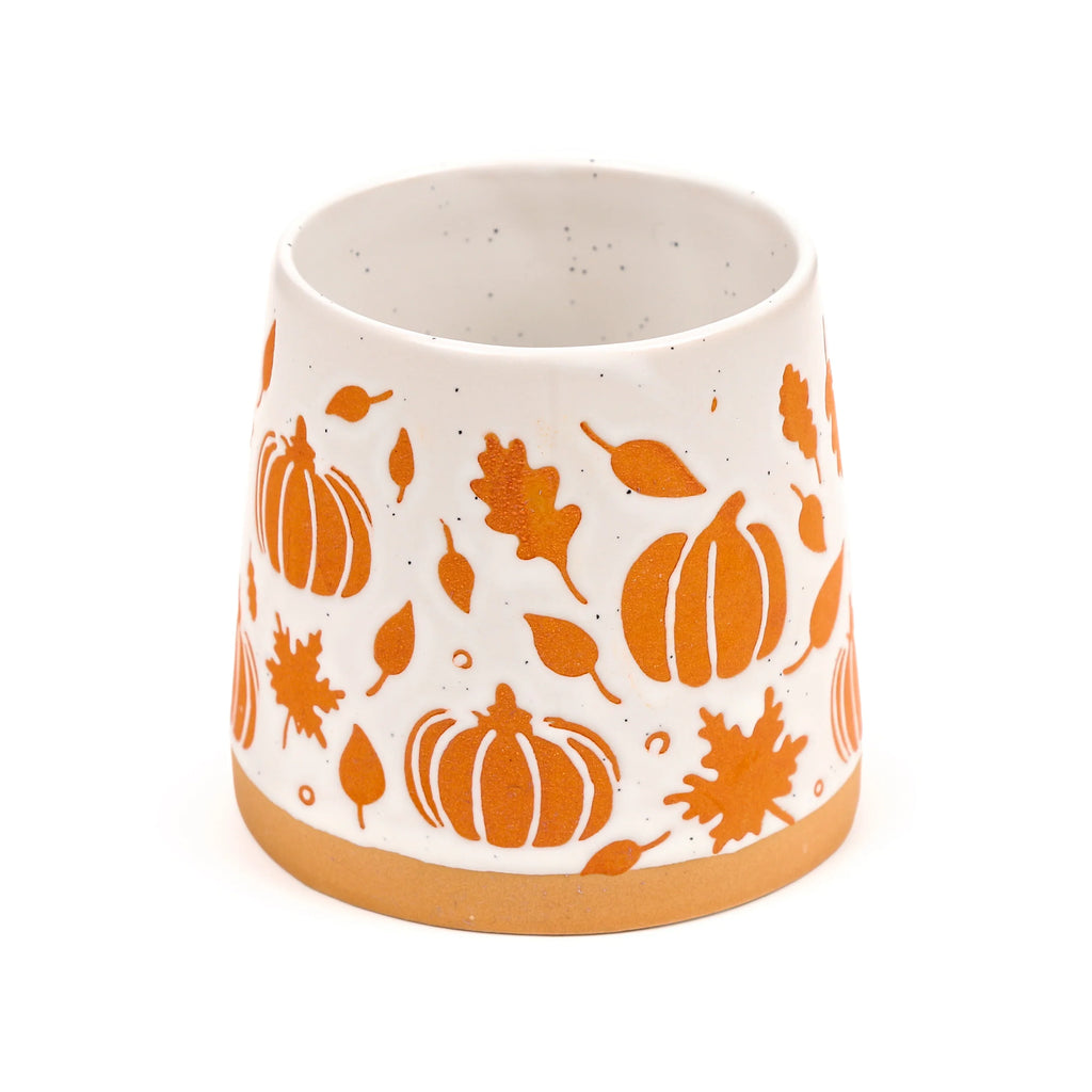 Angled view of a white speckled stoneware mug, featuring an embossed orange pumpkin and autumn leaf pattern, with a natural unglazed stoneware base.