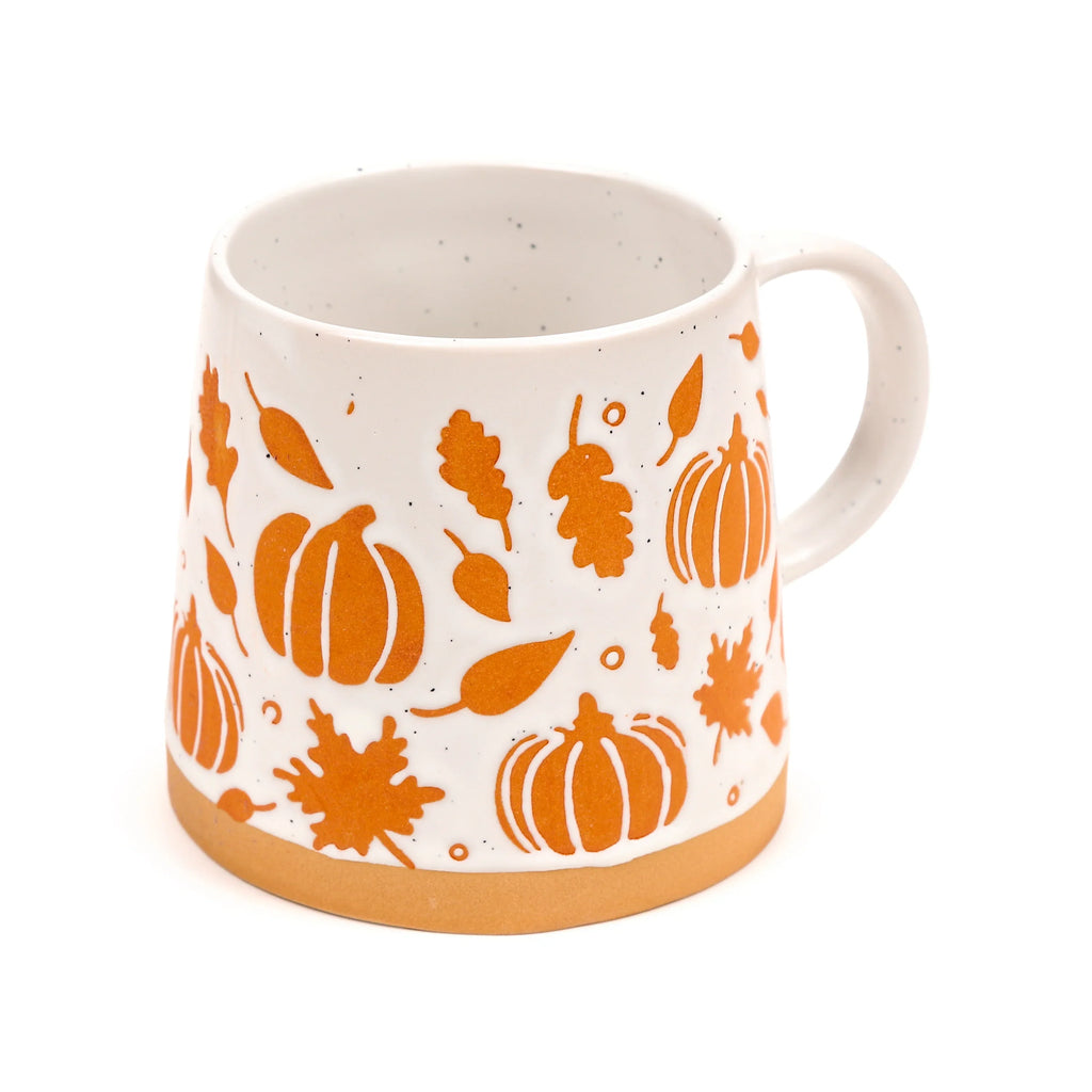 Angled view of a white stoneware mug, featuring orange pumpkin and autumn leaf patterns, a natural unglazed stoneware base, and subtle dark speckles on the white glaze.