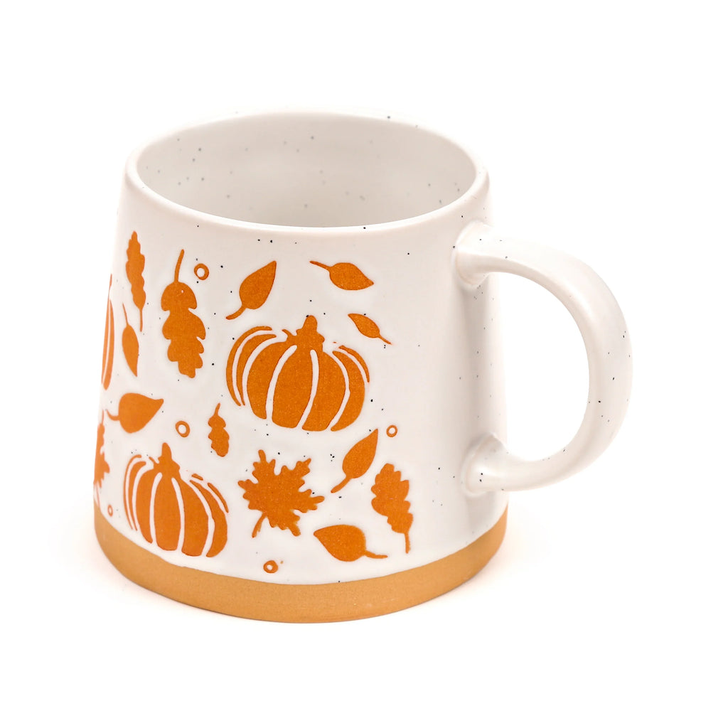 Angled view of a speckled off-white stoneware mug with an orange pumpkin and autumn leaf design, and an unglazed natural base.