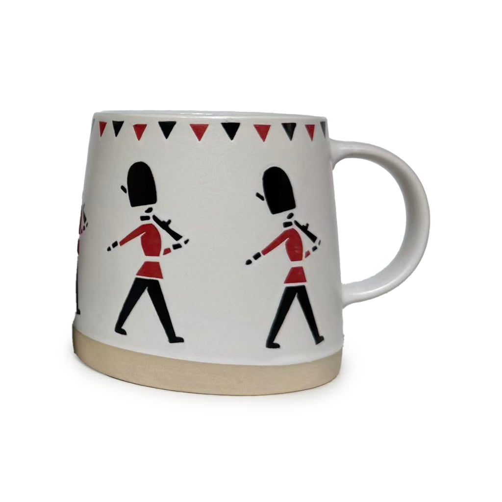 Angled view of a white tapered stoneware mug with a natural base, decorated with marching red and black Guardsmen figures and a triangular rim pattern, showing the handle.