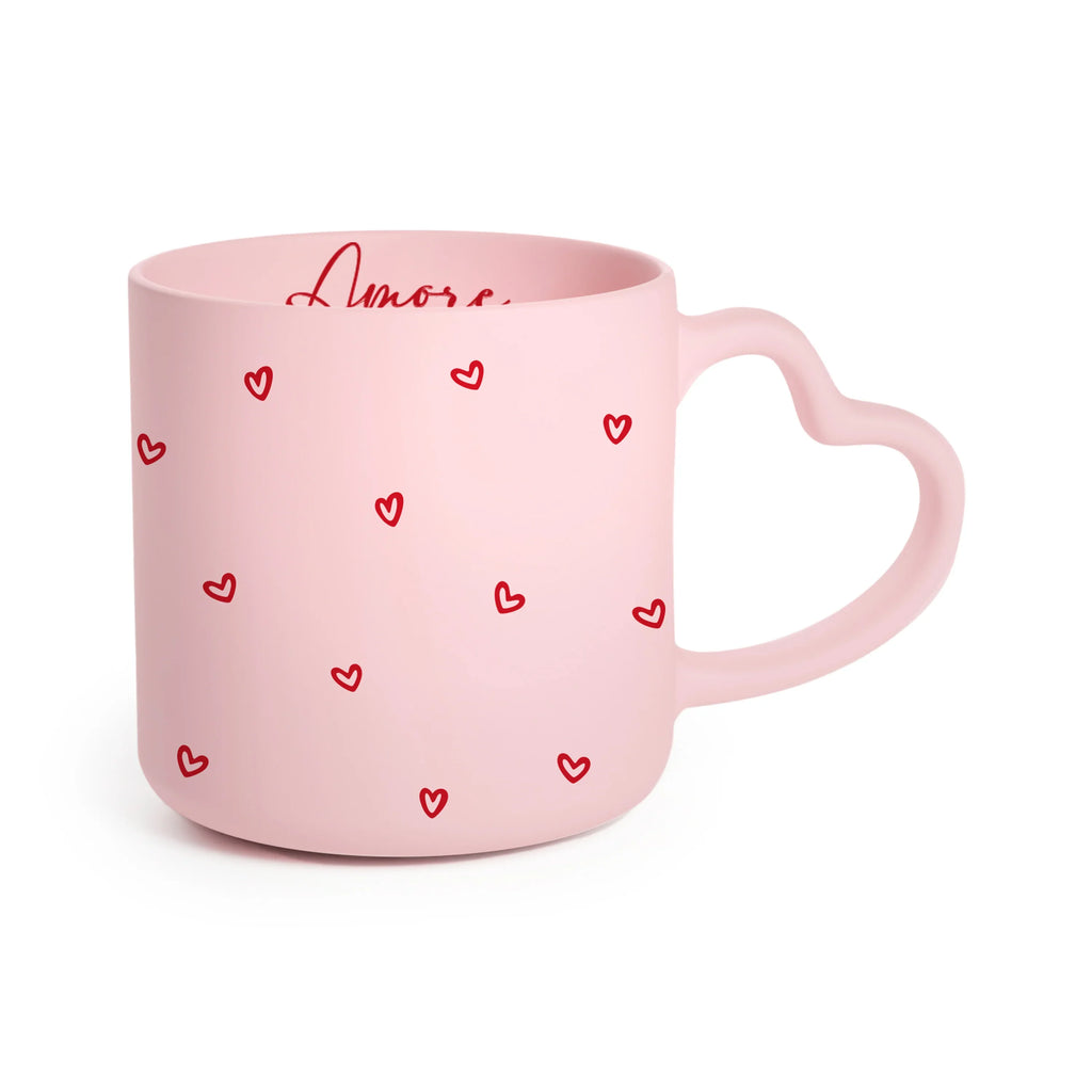 Angled view of a pink mug with a heart-shaped handle. The mug is decorated with scattered red heart patterns and the word 'Amore' in red script.