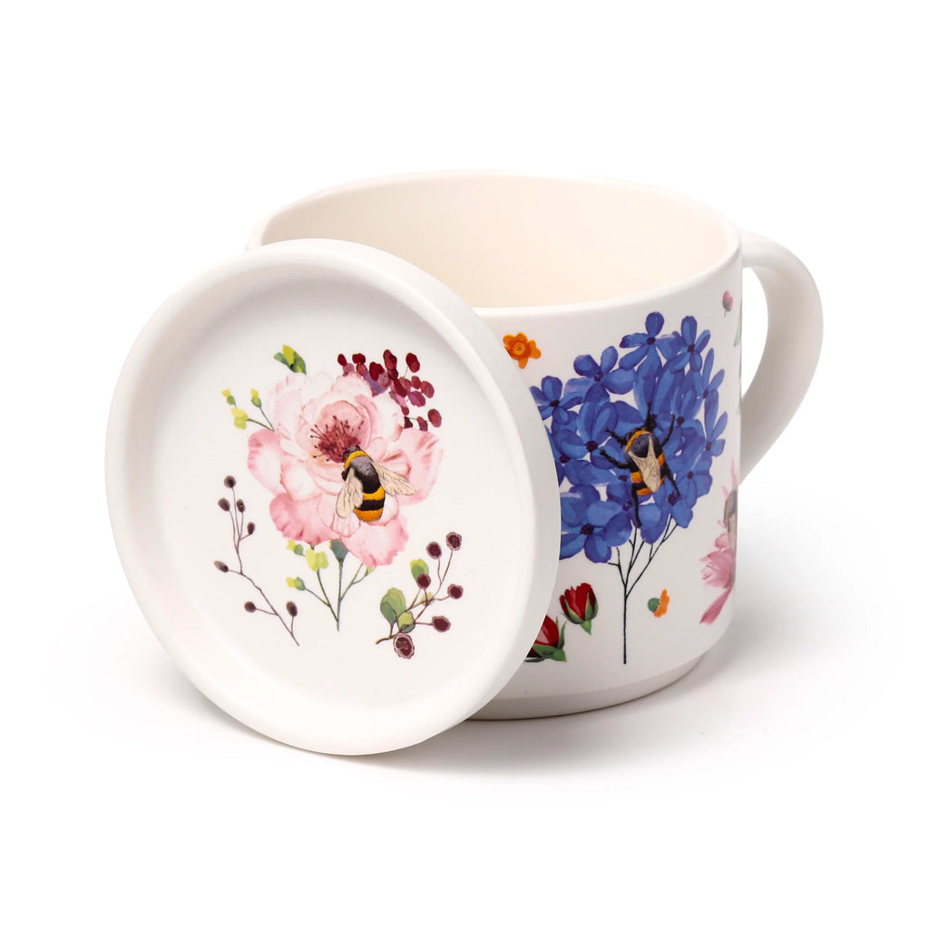 Angled view of a white porcelain mug and coaster lid set, featuring colourful floral and insect designs. The coaster lid displays a pink flower with a bee, whilst the mug shows blue flowers and a beetle.