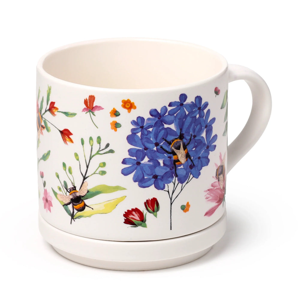 Angled view of a white porcelain mug featuring a colourful floral design with blue, red, and orange flowers, and bumblebee illustrations. The matching porcelain coaster lid is attached to the mug's base.