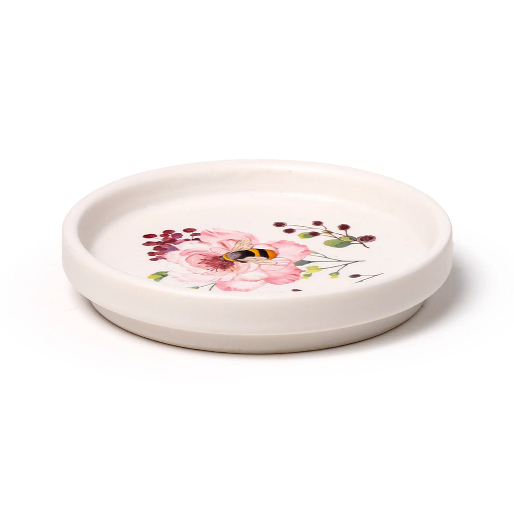 Angled view of a cream porcelain coaster lid, adorned with a pink floral and bumblebee illustration.