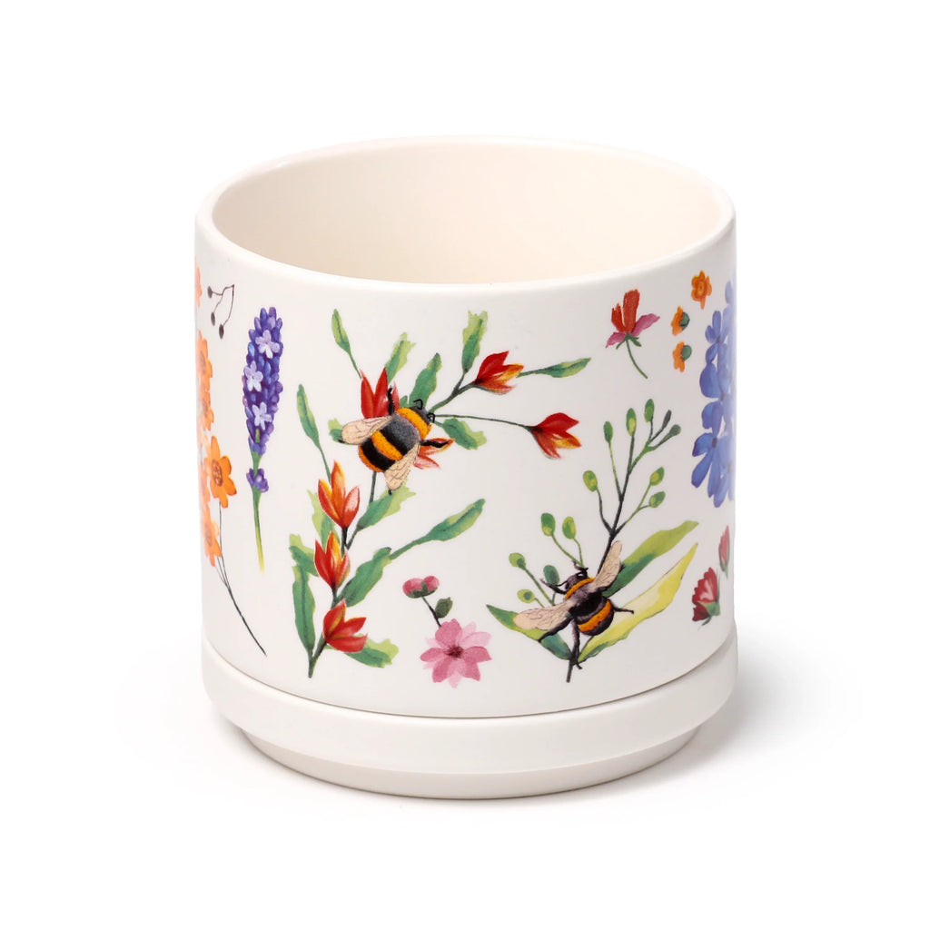 Angled view of a cream porcelain mug, featuring a vibrant, colourful floral and bee pattern in shades of red, orange, blue, purple, and pink, resting on its matching coaster lid.