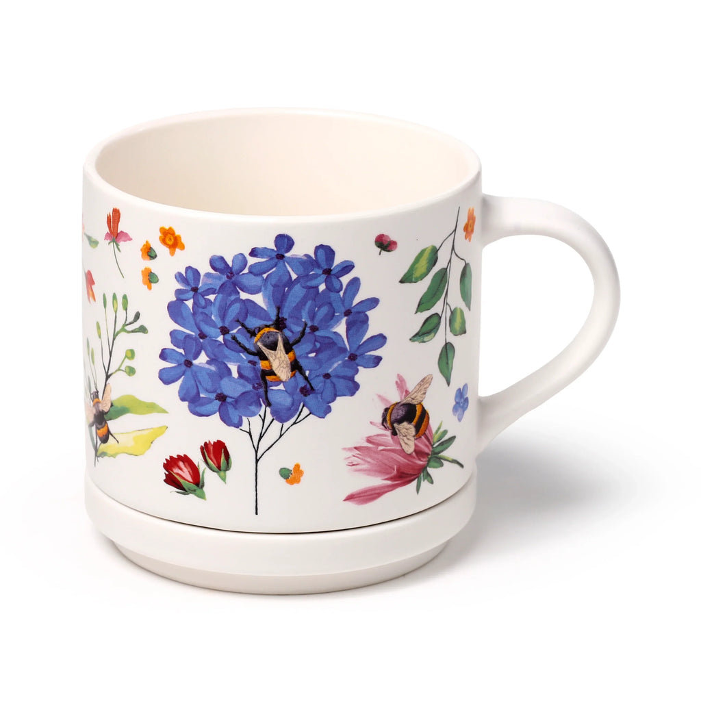 Angled view of a white porcelain mug, featuring a colourful Nectar Meadows design with blue flowers and bees, sitting in its matching coaster.
