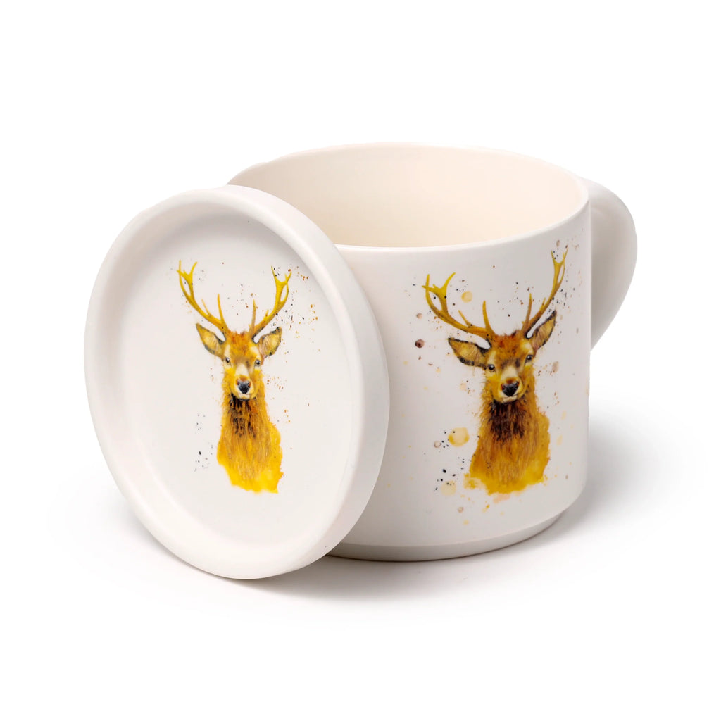 Angled view of a cream porcelain mug and matching coaster lid. Both items feature a vibrant watercolour-style illustration of a brown stag's head with prominent golden antlers and speckled detailing. The coaster lid is leaning beside the mug, showcasing its detailed stag design.