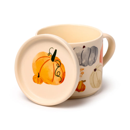Angled view of a cream porcelain mug and coaster lid set, featuring autumn harvest pumpkin and mushroom designs, with the lid displaying a large orange pumpkin and resting against the mug.