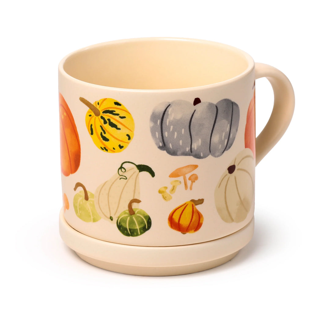 Angled view of a cream-coloured porcelain mug and coaster lid set, decorated with an autumn harvest pattern featuring various gourds and mushrooms.